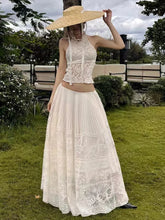 Woman in a white lace dress and wide-brimmed hat standing outdoors on grass with trees and benches in the background.
